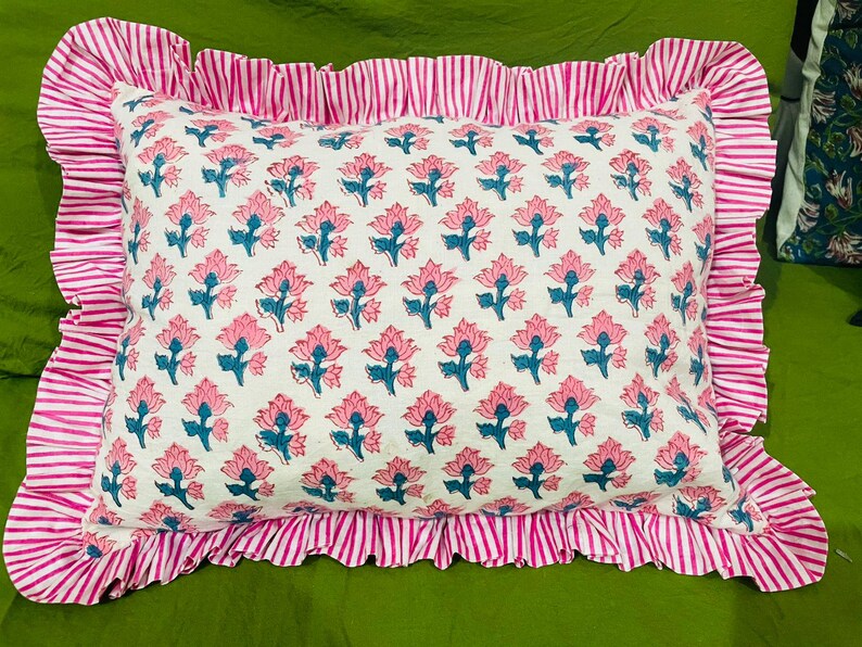 May include: A rectangular pillow featuring a white base with a repeating pattern of pink flowers and blue stems. The pillow is bordered by a wide, gathered ruffle trim in a pink and white vertical stripe pattern, creating a decorative edge.