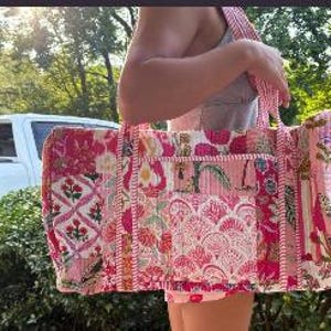 May include: A large, rectangular tote bag with a patchwork design in shades of pink and white. The bag features various floral and geometric patterns, with a striped strap and multiple pockets. The bag is being held by a person outdoors.