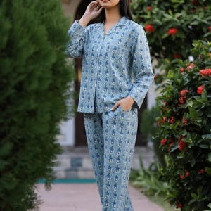 May include: A light blue pajama set featuring a repeating dark blue and white floral pattern. The set consists of a long-sleeved button-down shirt with a notched collar and a chest pocket, paired with matching loose-fitting trousers with side pockets.