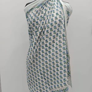 May include: A white sarong with a repeating pattern of blue floral designs and green leaves. The sarong is draped over a mannequin, showcasing its rectangular shape and decorative border. The fabric is lightweight and appears to be cotton.
