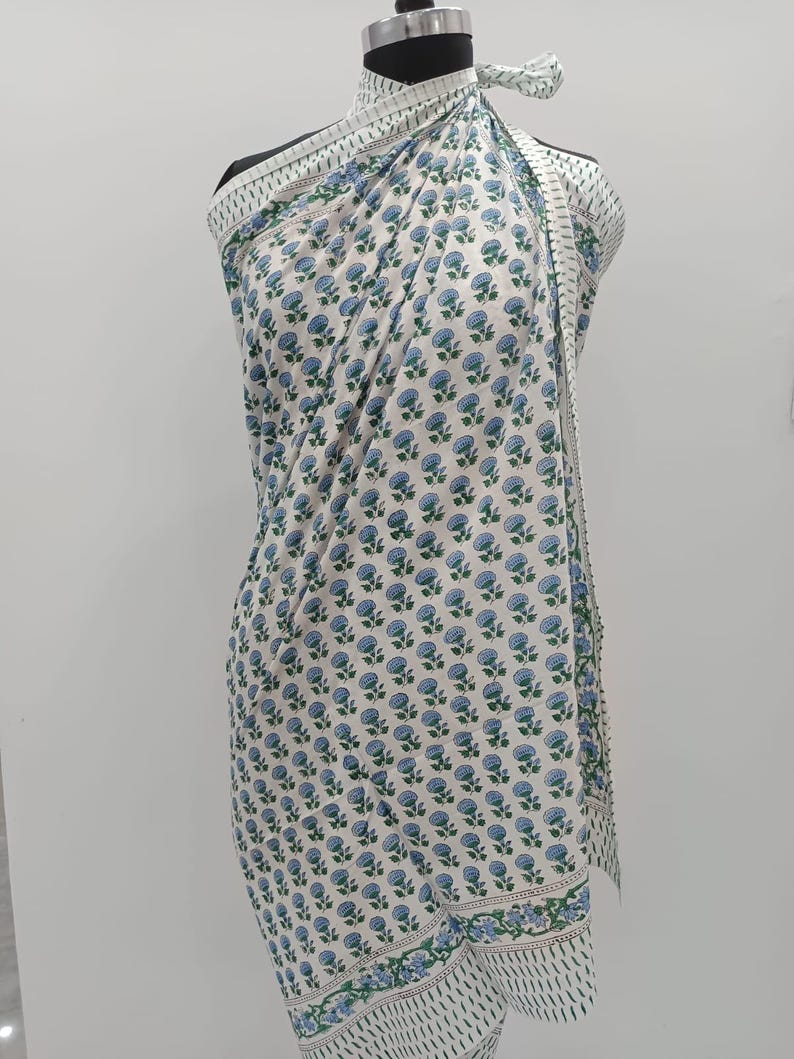May include: A white sarong with a repeating pattern of blue floral designs and green leaves. The sarong is draped over a mannequin, showcasing its rectangular shape and decorative border. The fabric is lightweight and appears to be cotton.
