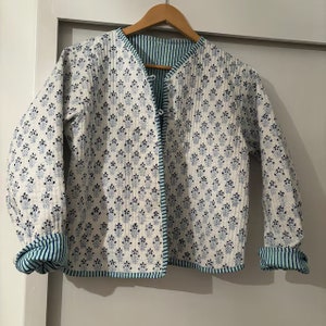 Peut inclure: Une veste matelassée blanche avec un motif floral bleu. La veste est doublée de rayures bleues et blanches et possède une fermeture à bouton bleu.