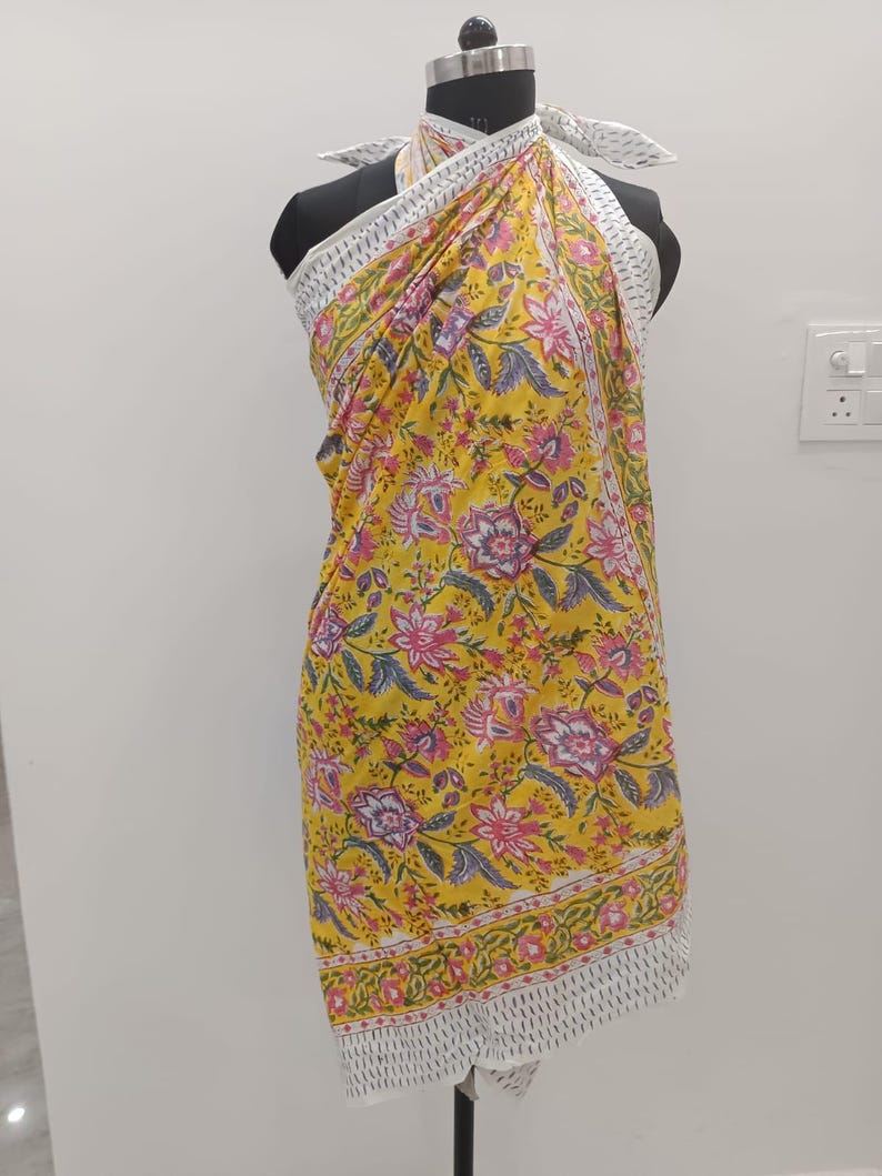 May include: A yellow sarong with a floral pattern in pink, purple, and green. The sarong has a white border with a geometric design. The fabric is draped over a mannequin, showcasing its design and style. This versatile accessory can be worn in multiple ways.