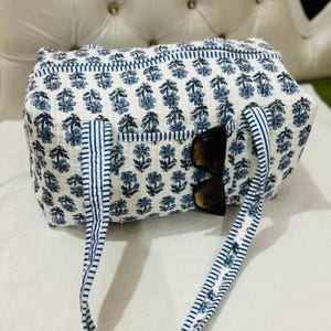 May include: A quilted duffel bag featuring a white base with a repeating blue floral block print pattern. The bag includes two blue and white striped fabric handles and a front exterior pocket with a pair of dark-tinted sunglasses tucked inside.