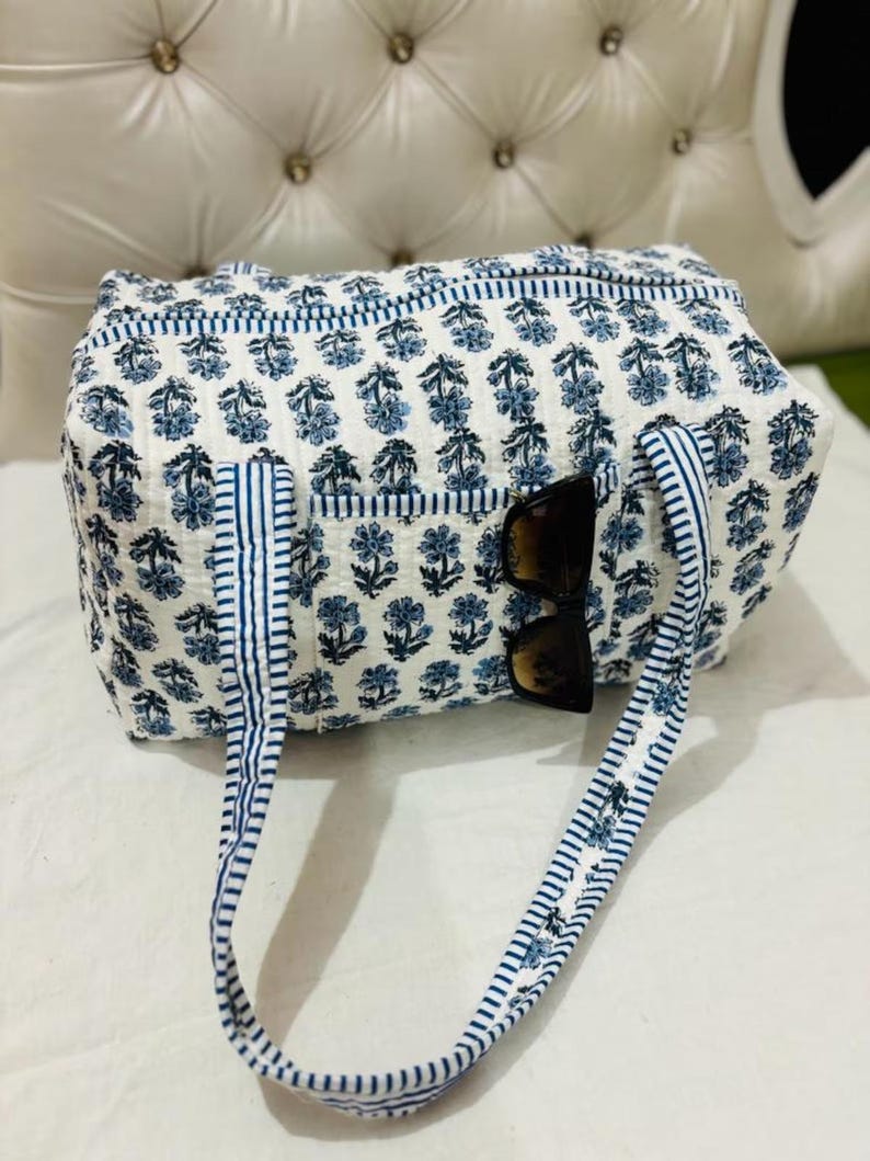 May include: A quilted duffel bag featuring a white base with a repeating blue floral block print pattern. The bag includes two blue and white striped fabric handles and a front exterior pocket with a pair of dark-tinted sunglasses tucked inside.
