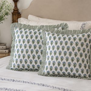 May include: Two square throw pillows featuring a white base with a repeating blue floral pattern and green stems. Each pillow is finished with a ruffled edge trim in a green and white striped fabric, creating a decorative border around the square shape.