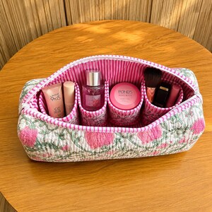 May include: A rectangular quilted cosmetic bag featuring a white exterior with a pink and green floral pattern. The interior is lined with pink and white vertical stripes and contains four cylindrical compartments holding various beauty products.