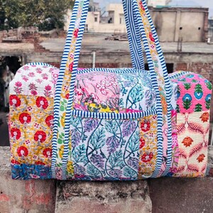 May include: A colorful patchwork duffel bag with floral and leaf patterns in pink, yellow, and blue. The bag has blue and white striped handles and trim. The bag is made of quilted fabric.