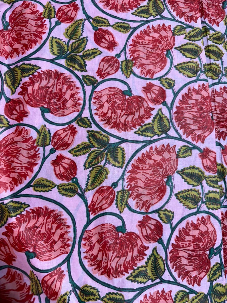 Pu&ograve; includere: Primo piano di un motivo floreale con grandi fiori e boccioli rossi, foglie e steli verdi su sfondo rosa chiaro. Il disegno si ripete sul tessuto, creando un tessuto visivamente accattivante.