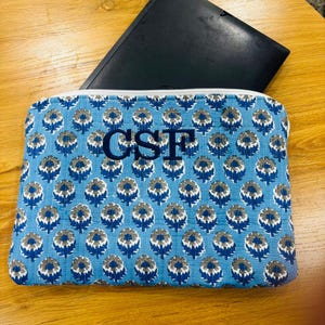 May include: A blue floral patterned laptop sleeve with the initials "CSF" embroidered in navy blue. The sleeve is made of quilted fabric and has a white zipper. A black laptop is partially visible.
