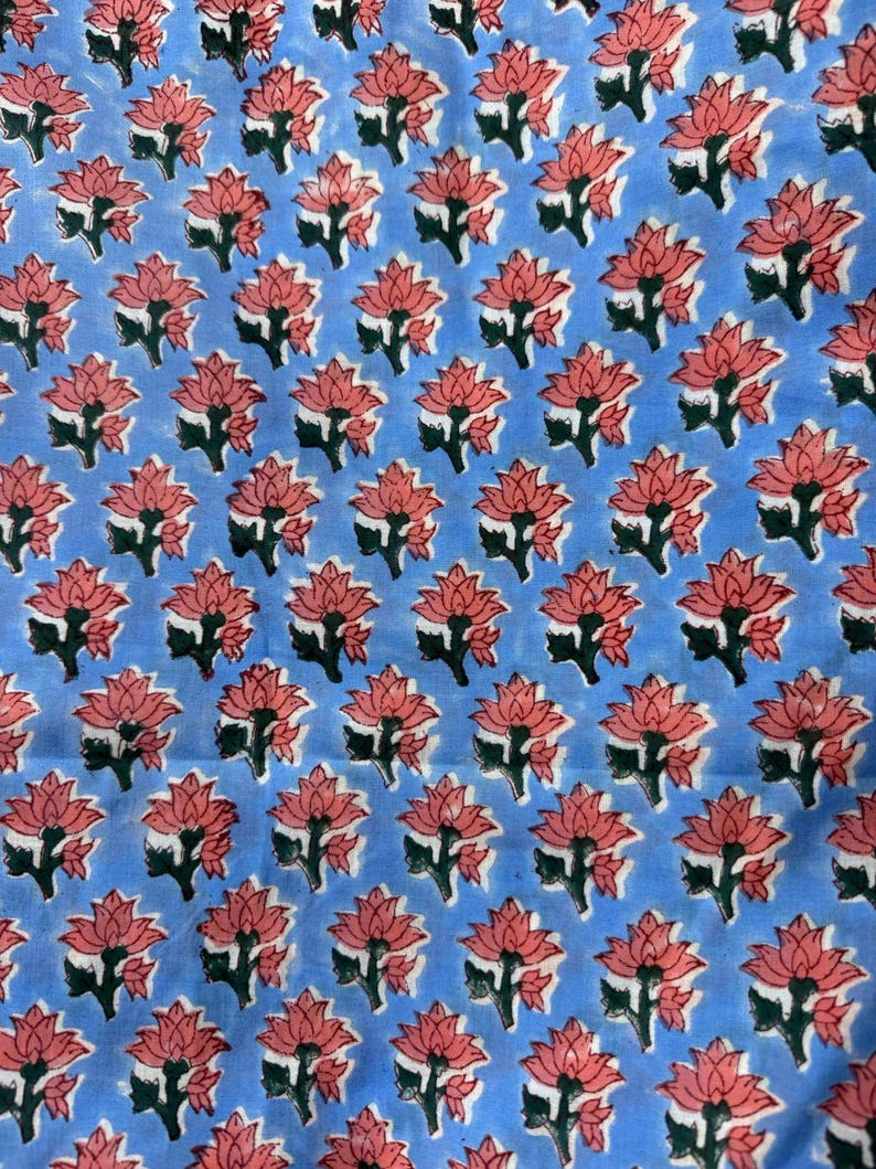 May include: A light blue fabric with a repeating pattern of pink lotus flowers and green leaves. The flowers are evenly spaced across the fabric, creating a visually appealing design. The fabric appears to be cotton or a similar material.