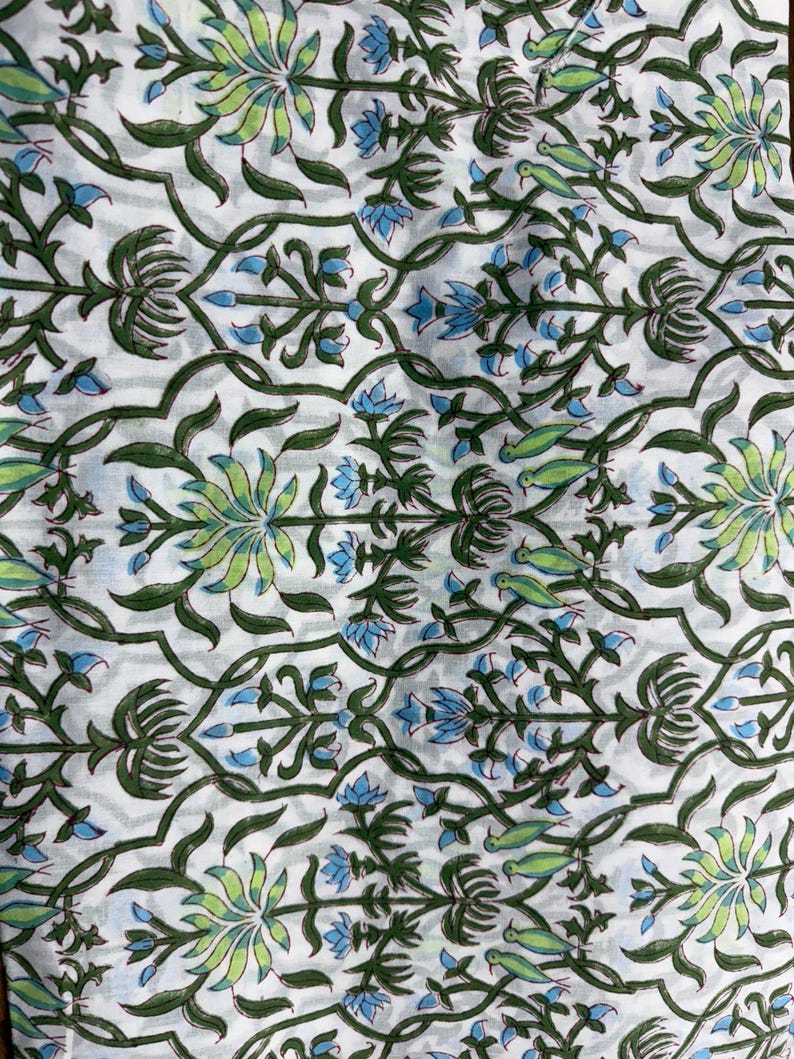 May include: White fabric with a repeating floral pattern in green, light green, and blue. The design features stylized flowers, leaves, and bird-like shapes, creating a botanical theme. The fabric has a soft, slightly wrinkled appearance.