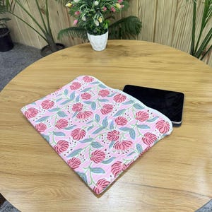 May include: A folded pink floral patterned fabric with red flowers and green leaves. The fabric is on a wooden table next to a black electronic device. A small potted plant is in the background.