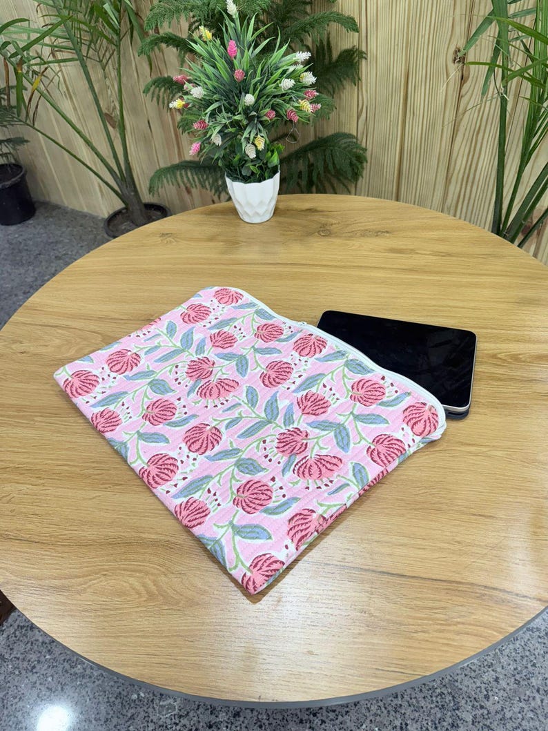 May include: A folded pink floral patterned fabric with red flowers and green leaves. The fabric is on a wooden table next to a black electronic device. A small potted plant is in the background.