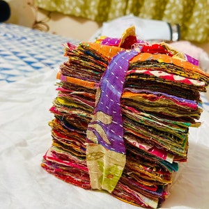 May include: A stack of square fabric scraps in various colors including orange, purple, red, and yellow, bound together by a purple and green patterned fabric strip. Each piece of fabric features visible white running stitch embroidery details.