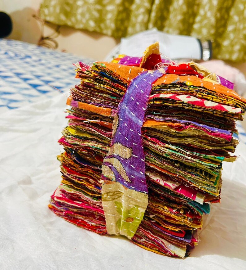 May include: A stack of square fabric scraps in various colors including orange, purple, red, and yellow, bound together by a purple and green patterned fabric strip. Each piece of fabric features visible white running stitch embroidery details.
