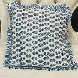 Op de afbeelding: Een vierkant sierkussen met een witte stoffen basis en een herhalend blauw bloemenpatroon in blokdruk. Het kussen is afgewerkt met een geplooide rand van blauw en wit gestreepte stof die alle vier de zijden van het kussen omlijst.