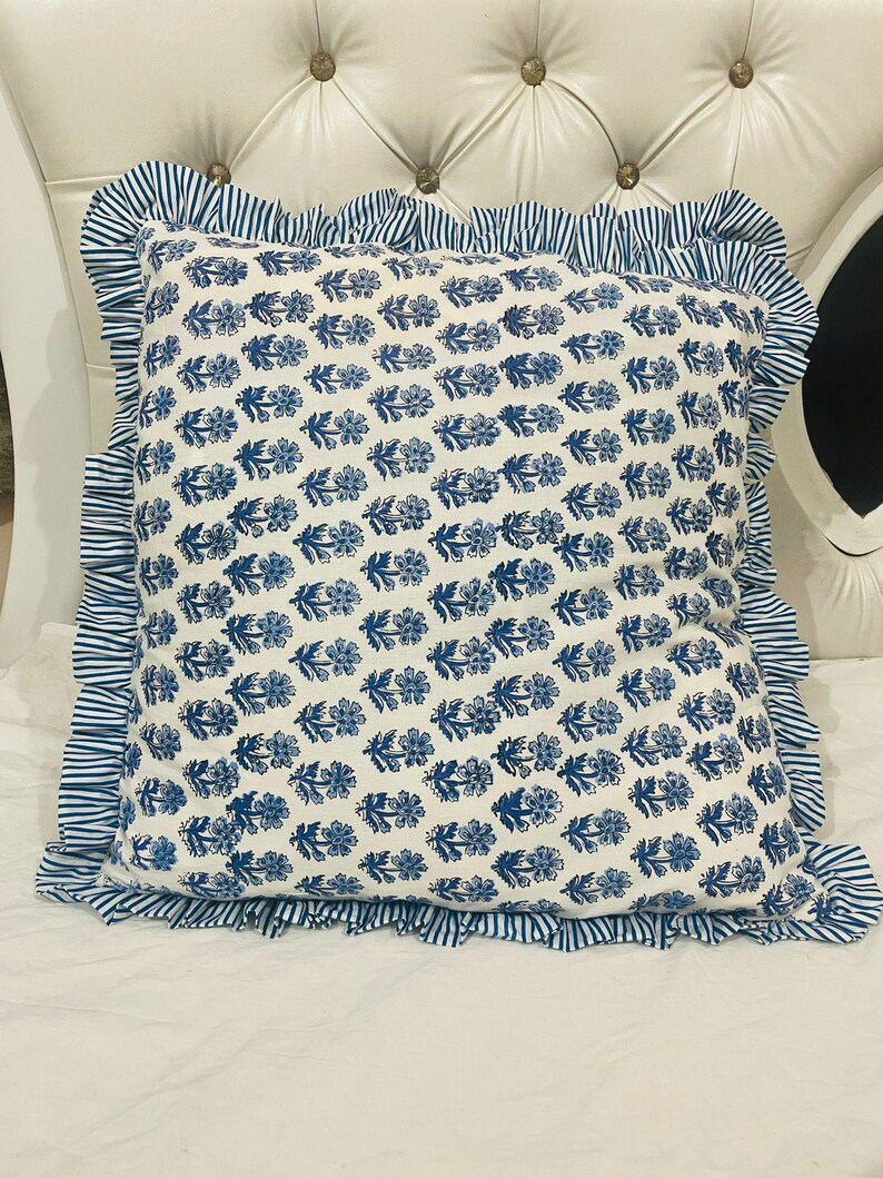 Op de afbeelding: Een vierkant sierkussen met een witte stoffen basis en een herhalend blauw bloemenpatroon in blokdruk. Het kussen is afgewerkt met een geplooide rand van blauw en wit gestreepte stof die alle vier de zijden van het kussen omlijst.