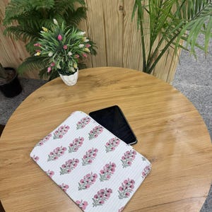May include: White floral patterned laptop sleeve with pink and green accents. The sleeve is rectangular and features a repeating pattern of stylized flowers. A black smartphone rests on the wooden table next to the sleeve.