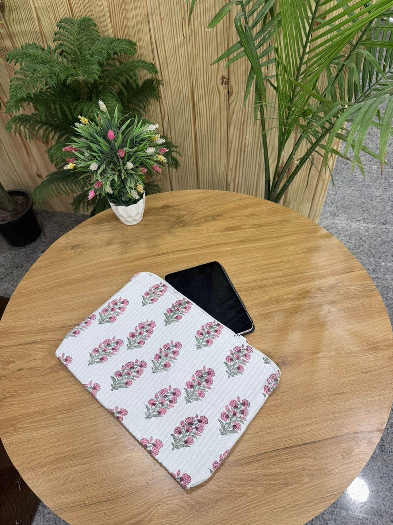May include: White floral patterned laptop sleeve with pink and green accents. The sleeve is rectangular and features a repeating pattern of stylized flowers. A black smartphone rests on the wooden table next to the sleeve.