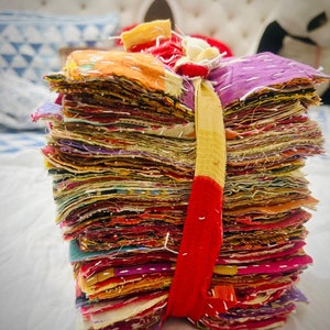 May include: A tall, vertical stack of numerous square fabric scraps in a wide variety of vibrant colors including purple, orange, red, and yellow. The fabric pieces are held together by a single, long, thin strip of yellow and red fabric tied around them.