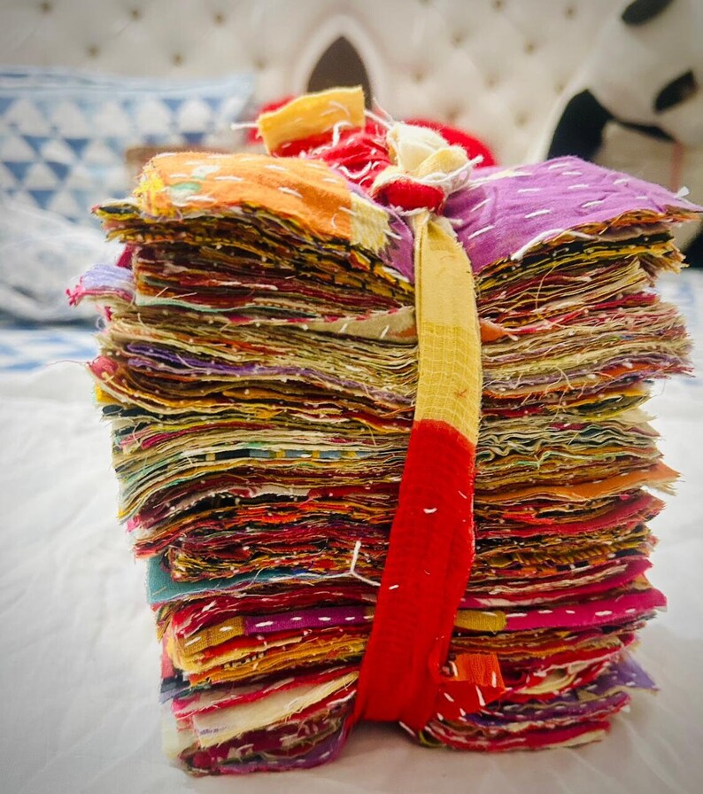 May include: A tall, vertical stack of numerous square fabric scraps in a wide variety of vibrant colors including purple, orange, red, and yellow. The fabric pieces are held together by a single, long, thin strip of yellow and red fabric tied around them.