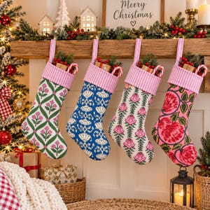 Op de afbeelding: Vier kerstsokken hangen op een rij, elk met een roze en wit gestreepte boord en een uniek bloemenpatroon. De sokken tonen groene, blauwe, crèmekleurige en roze stoffen ontwerpen, met kleine cadeautjes en zuurstokken erin gestoken.