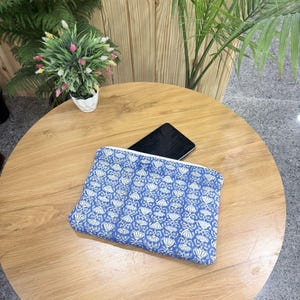 May include: A blue and white patterned clutch bag with a zipper closure. The bag is rectangular and lies on a wooden table. A black tablet is partially visible inside the bag. Decorative plants are in the background.