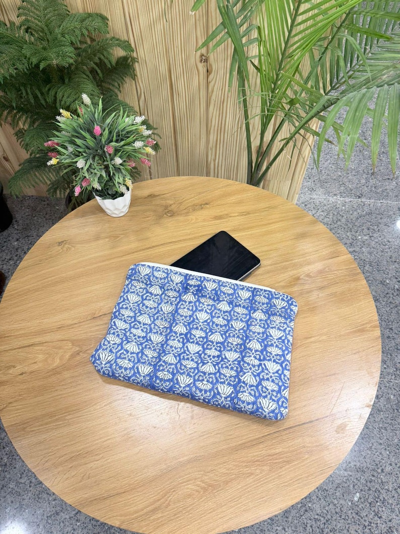 May include: A blue and white patterned clutch bag with a zipper closure. The bag is rectangular and lies on a wooden table. A black tablet is partially visible inside the bag. Decorative plants are in the background.