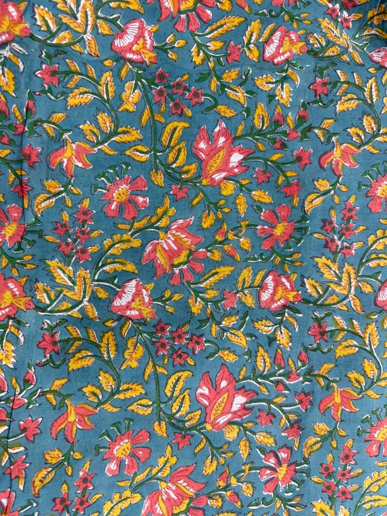 Pu&ograve; includere: Primo piano di un tessuto turchese con un motivo floreale. Il design presenta fiori rossi e gialli con foglie e viti verdi. I fiori hanno accenti bianchi e le foglie sono di un giallo brillante.