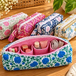 May include: A collection of five rectangular quilted fabric pouches featuring floral patterns in pink, magenta, navy blue, and yellow. One open pouch in the foreground displays a pink and white gingham interior lining with multiple internal compartments.