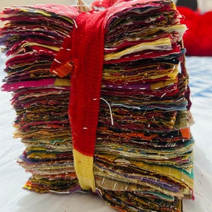 May include: A tall stack of square fabric scraps featuring various colors and patterns, bound together by a red and yellow fabric strip. Each piece of fabric displays visible white running stitch embroidery details along the edges and surfaces.