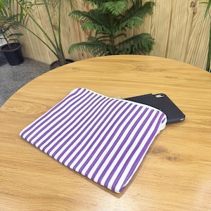 May include: A white and purple striped pouch with a zipper closure rests on a wooden table. A black smartphone is partially visible behind the pouch. The background features green plants.