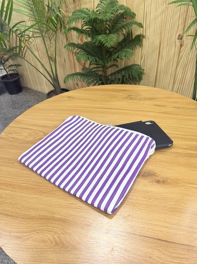 May include: A white and purple striped pouch with a zipper closure rests on a wooden table. A black smartphone is partially visible behind the pouch. The background features green plants.