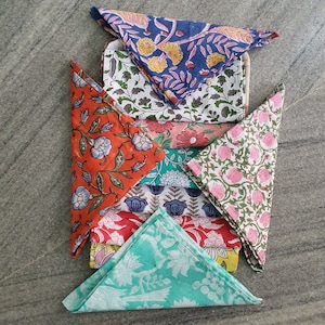 May include: A collection of folded square napkins featuring various block-printed floral and botanical patterns in vibrant colors including orange, blue, pink, green, and yellow. The fabric pieces are stacked and arranged to display their designs.