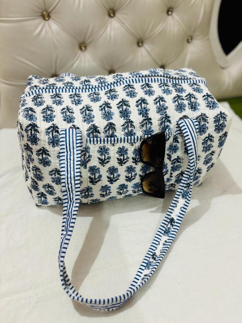 May include: A white tote bag with a blue floral pattern and blue striped handles. The bag has a pocket on the front and sunglasses are tucked inside. The bag is made of a quilted fabric.