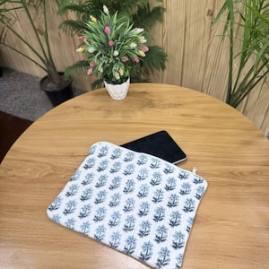 May include: A white and blue floral pattern laptop sleeve with a zipper closure. The sleeve is on a wooden table, with a black tablet partially visible inside. Decorative plants are in the background.