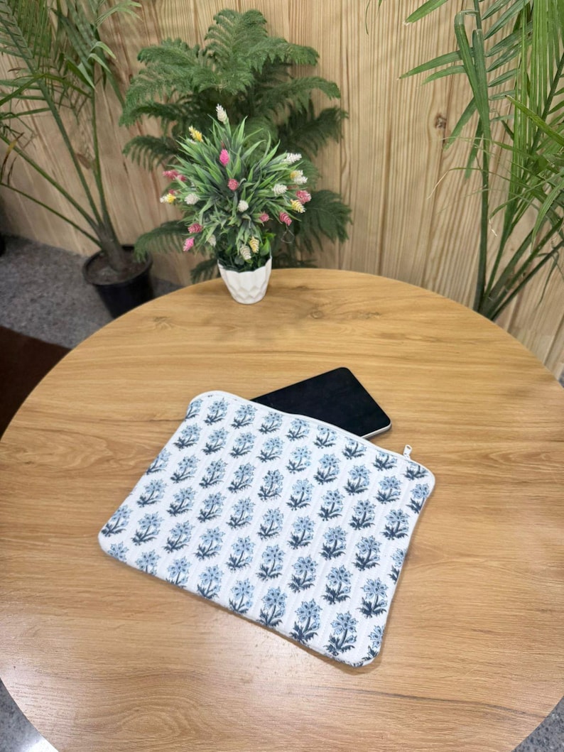 May include: A white and blue floral pattern laptop sleeve with a zipper closure. The sleeve is on a wooden table, with a black tablet partially visible inside. Decorative plants are in the background.