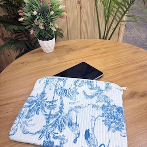 May include: White and blue toiletry bag with a zipper closure. The bag features a blue botanical and animal print on a white quilted background. A black smartphone rests on the wooden table behind the bag.