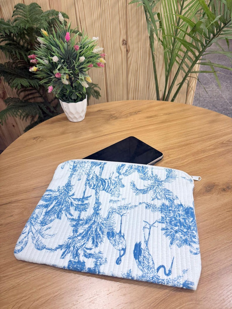 May include: White and blue toiletry bag with a zipper closure. The bag features a blue botanical and animal print on a white quilted background. A black smartphone rests on the wooden table behind the bag.