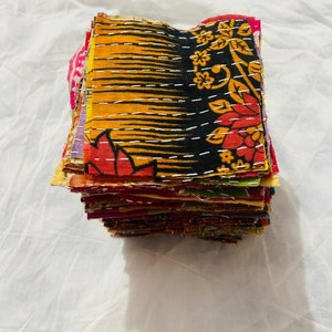 May include: A stack of square fabric patches featuring various colorful patterns, including floral and geometric designs in black, orange, red, and yellow. Each piece of fabric is held together with visible white running stitch embroidery.