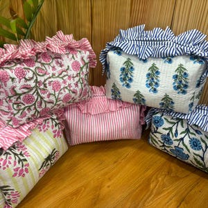 Puede incluir: Una colección de cinco estuches rectangulares acolchados con bordes de volantes. Los estuches presentan varios patrones, incluyendo estampados florales rosas, motivos florales azules y rayas rosas y blancas, con volantes a juego.