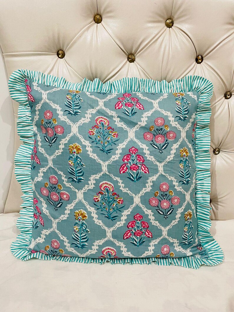 May include: A square throw pillow featuring a teal blue background with a white lattice pattern and repeating floral motifs in pink and yellow. The pillow is finished with a ruffled border in a teal and white striped fabric along all four edges.
