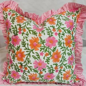 May include: A square throw pillow featuring a white base with a repeating floral pattern in shades of pink and orange, accented by green vines. The pillow is finished with a ruffled border made of fabric featuring thin pink and white stripes.