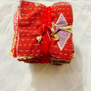 May include: A stack of square fabric swatches tied together with a red fabric strip. The top swatch is primarily red with white running stitch embroidery and features a light purple diamond pattern. The edges of the fabric pieces are frayed.