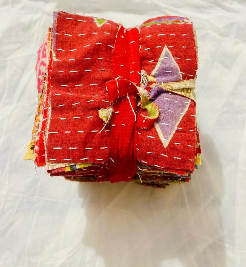 May include: A stack of square fabric swatches tied together with a red fabric strip. The top swatch is primarily red with white running stitch embroidery and features a light purple diamond pattern. The edges of the fabric pieces are frayed.