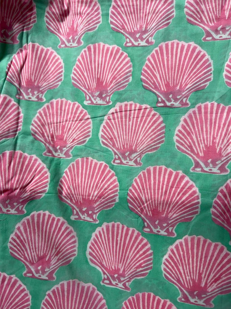 Pu&ograve; includere: Un motivo ripetuto di conchiglie rosa con strisce bianche su uno sfondo verde acqua. Le conchiglie sono uniformemente distanziate e creano un design estivo a tema spiaggia. L'immagine &egrave; un primo piano del tessuto.