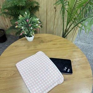 May include: A pink and white gingham patterned zippered pouch rests on a wooden table next to a black tablet. The pouch is made of a soft fabric. The table is surrounded by plants.