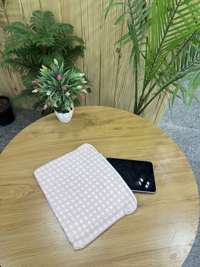 May include: A pink and white gingham patterned zippered pouch rests on a wooden table next to a black tablet. The pouch is made of a soft fabric. The table is surrounded by plants.