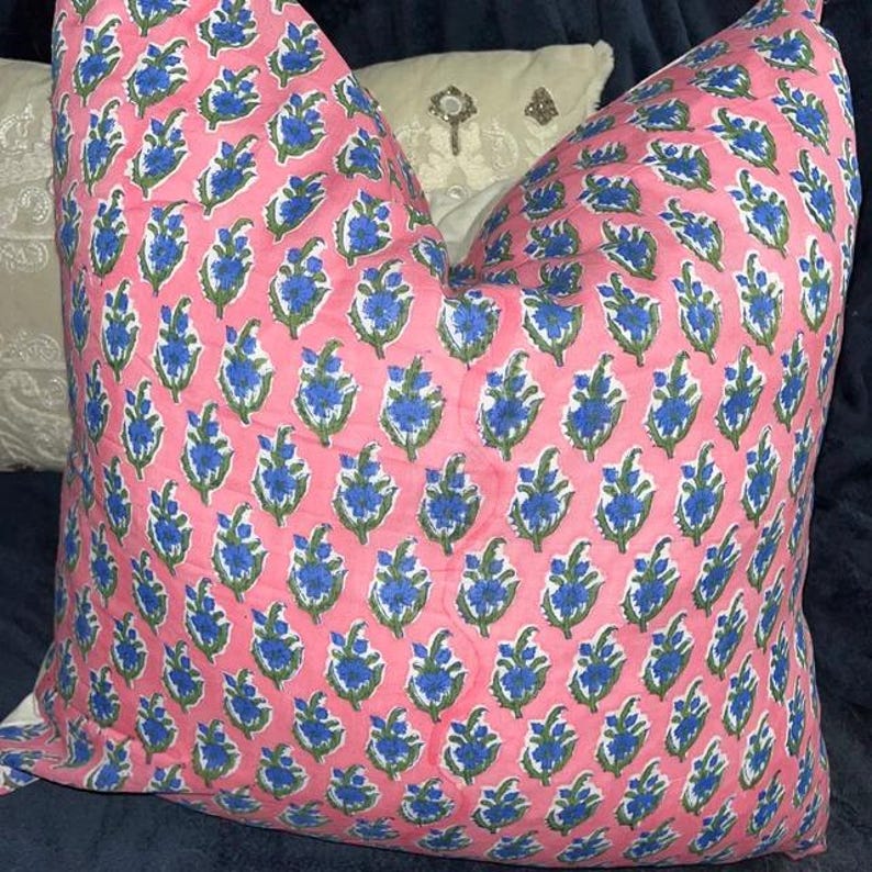 Puede incluir: Un coj&iacute;n cuadrado con un patr&oacute;n floral repetitivo. La tela es de un color rosa suave con un motivo recurrente de flores azules y hojas verdes dispuestas en una cuadr&iacute;cula vertical. El coj&iacute;n tiene una apariencia mullida, suave y cuadrada.